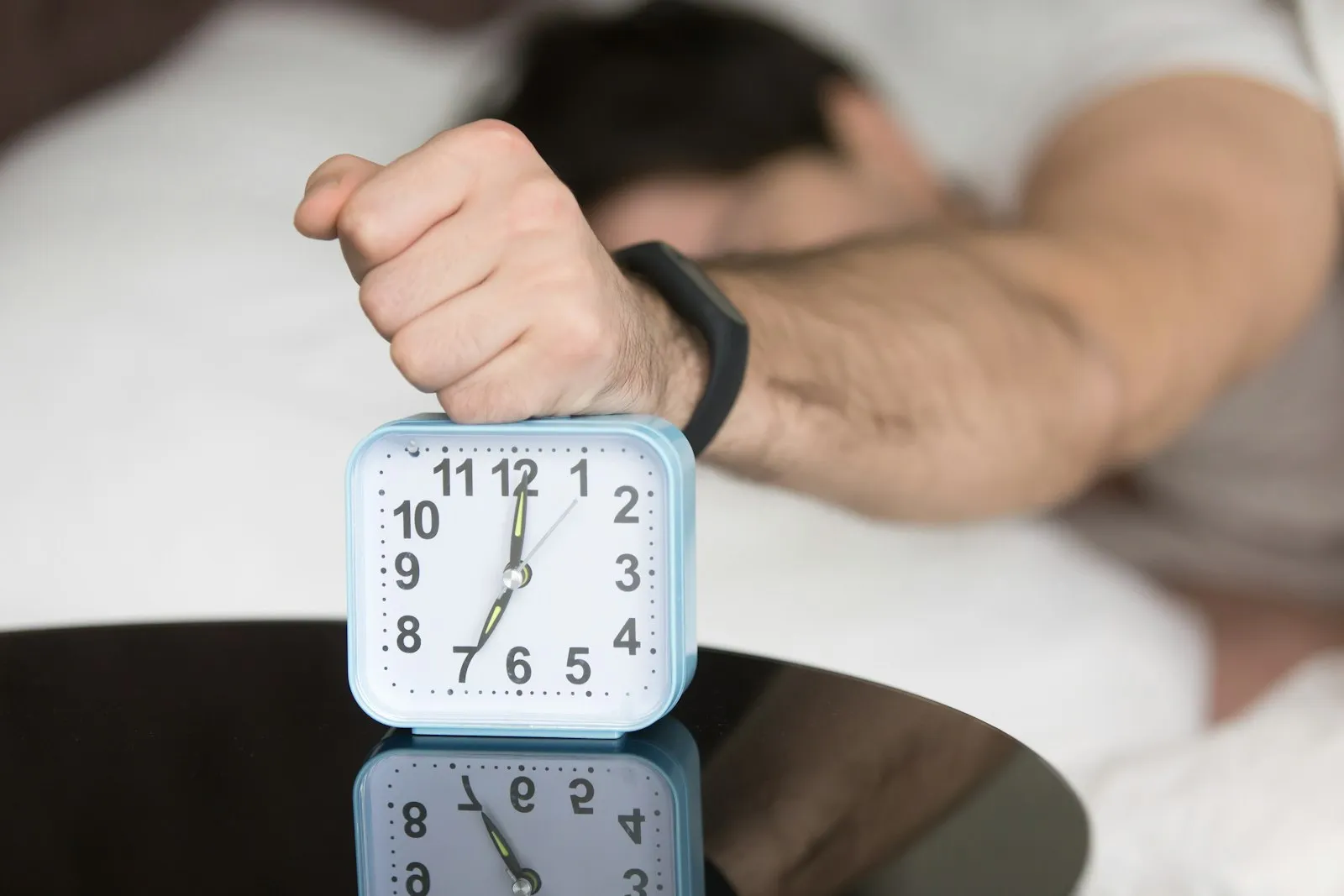 A man lying in bed, slamming his fist down on a blue alarm clock